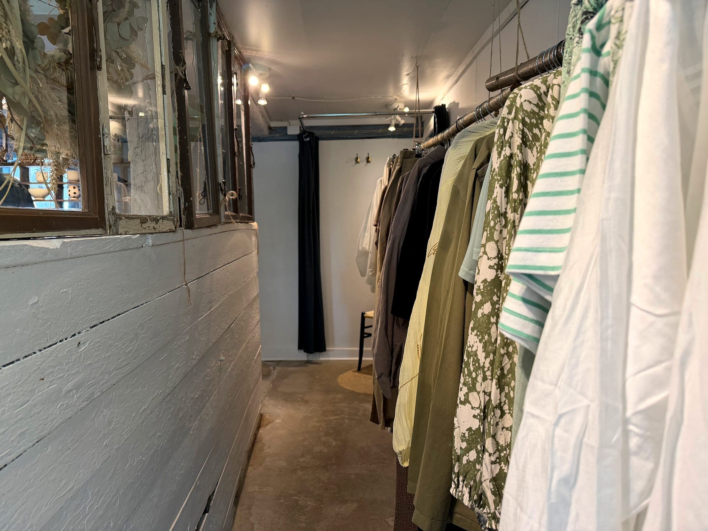 Schmaler Gang im Laden mit Kleidung auf Ständern und Trockenblumen im Fenster